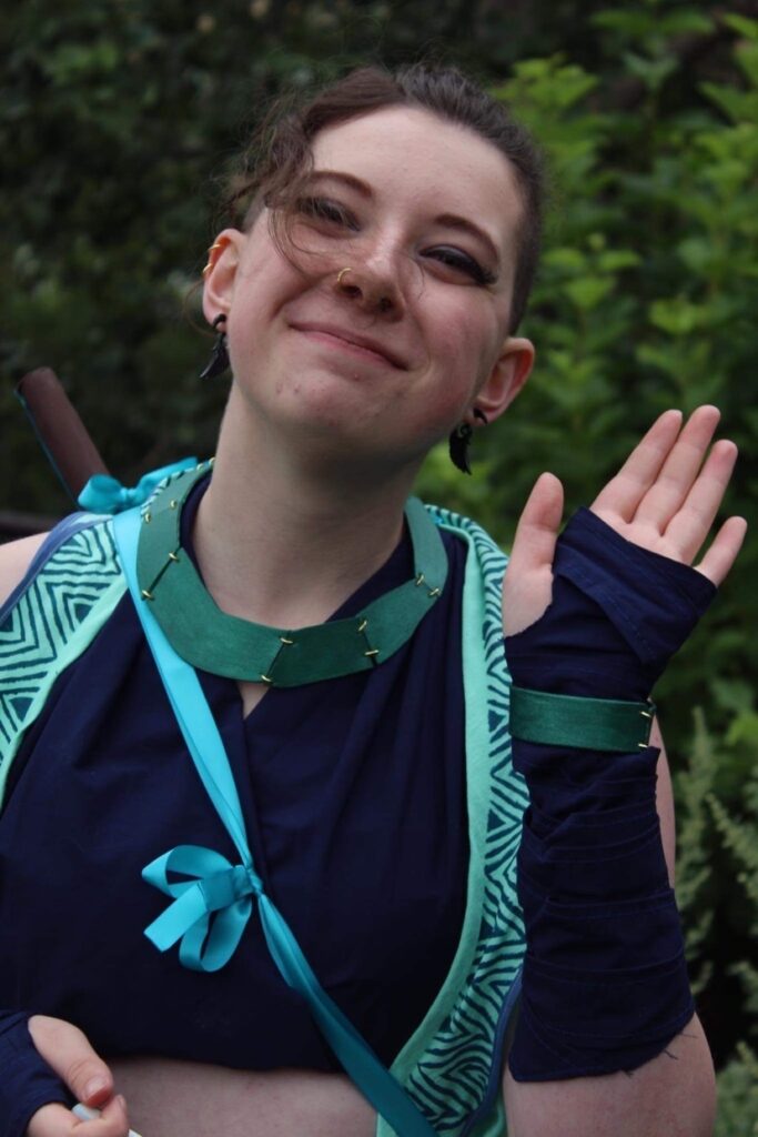 A headshot of Molly in their Beauregard cosplay. They are waving.